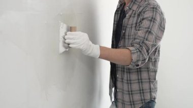 Eldiven ve ekose gömlek giyen tanınmamış inşaat işçisi, bir ev yenileme veya inşaat projesi sırasında beyaz bir duvara sıva uygulamak için macun bıçağı kullanıyor..