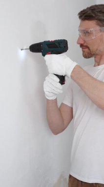 Beyaz tişört ve koruyucu eldiven giyen orta yaşlı erkek işçi, ev yenileme çalışmaları sırasında kablosuz matkapla duvarda delik açıyor. Dikey görünüm.