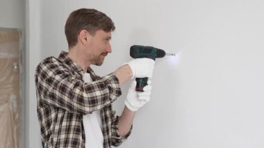 İşaretli gömlek ve beyaz eldiven giyen orta yaşlı bir adam, ev yenileme çalışmaları sırasında kablosuz matkapla duvarda delik açıyor. Dikey görünüm.