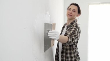 Kareli gömlek giyen kadın inşaat işçisi, beyaz duvar yüzeyini düzleştiriyor, profesyonel spatula tekniği ile macunu uyguluyor, iç dekorasyon konusunda yetenekli..