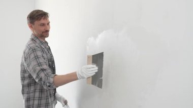 Koruyucu eldiven ve ekose gömlek giyen erkek inşaat işçisi, tadilat projesi sırasında profesyonel alçı levha bantlama bıçağı kullanarak duvarın yüzeyine sorunsuzca alçı yayıyor. Dikey görünüm.