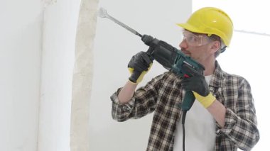 Bej kareli gömlek, sarı başlık ve koruyucu eldiven giyen inşaat işçisi, döner çekiç matkabıyla beyaz duvarı yıkıyor, toz ve portre görüntüsü yaratıyor. Yenileme konsepti.