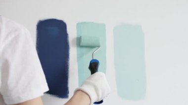 Eldiven giyen ressam beyaz bir duvara boya silindiri uyguluyor, mavi rengin çeşitli tonlarını test ederek iç tasarım için güzel bir eğim etkisi yaratıyor.