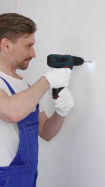 Mavi üniformalı profesyonel işçi, güvenlik eldivenleri, tadilat sırasında beyaz duvarda delik açmak için kablosuz matkap kullanıyor. Dikey portre görünümü.