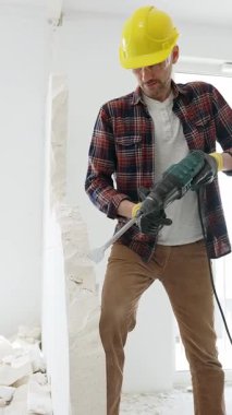 Bej kareli gömlek, sarı başlık ve koruyucu eldiven giyen inşaat işçisi, dönen çekiç matkabıyla beyaz duvarı yıkıyor ve orta uzunlukta toz üretiyor. Yenileme konsepti
