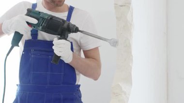 Mavi tulumlu ve koruyucu beyaz eldivenli tanımlanamayan erkek inşaat işçisi, beyaz duvarı döner çekiç matkapıyla yıkıyor, toz üretiyor ve yatay görüşü kapatıyor. Yenileme