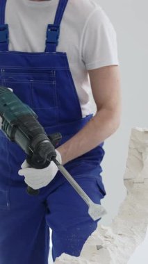 Mavi tulumlu ve koruyucu beyaz eldivenli tanımlanamayan erkek inşaat işçisi, beyaz duvarı döner çekiç matkapıyla yıkıyor, toz üretiyor ve yatay görüşü kapatıyor. Yenileme