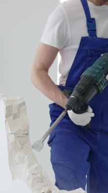 Mavi tulumlu ve koruyucu beyaz eldivenli tanımlanamayan erkek inşaat işçisi, beyaz duvarı döner çekiç matkapıyla yıkıyor, toz üretiyor ve yatay görüşü kapatıyor. Yenileme