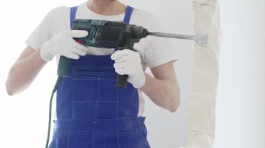 Mavi tulumlu ve koruyucu beyaz eldivenli tanımlanamayan erkek inşaat işçisi, beyaz duvarı döner çekiç matkapıyla yıkıyor, toz üretiyor ve yatay görüşü kapatıyor. Yenileme