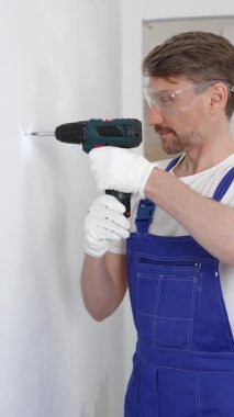 Mavi üniformalı profesyonel işçi, güvenlik eldivenleri, tadilat sırasında beyaz duvarda delik açmak için kablosuz matkap kullanıyor. Dikey portre görünümü.