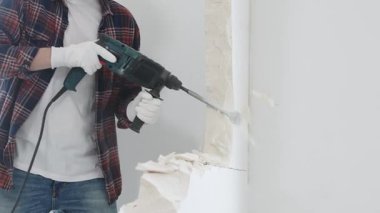 Kırmızı kareli kot pantolon ve koruyucu eldiven giyen kimliği belirsiz erkek inşaat işçisi, döner çekiç matkabıyla beyaz duvarı yıkıyor, toz üretiyor, yakın görüş sağlıyor. Yenileme konsepti.