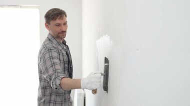 Eldiven ve ekose gömlek giyen orta yaşlı inşaat işçisi, yenileme projesi sırasında pürüzsüz bir yüzey oluşturmak için beyaz duvar alçılamak için son malayı kullanıyor..