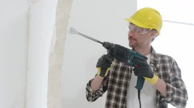 Bej kareli gömlek, sarı başlık ve koruyucu eldiven giyen inşaat işçisi, döner çekiç matkabıyla beyaz duvarı yıkıyor, toz ve portre görüntüsü yaratıyor. Yenileme konsepti.