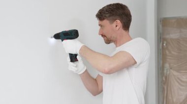 Beyaz tişört ve koruyucu eldiven giyen orta yaşlı erkek işçi, ev yenileme çalışmaları sırasında kablosuz matkapla duvarda delik açıyor. Dikey görünüm.