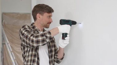 İşaretli gömlek ve güvenlik eldiveni giyen erkek inşaat işçisi, kablosuz tornavida kullanarak duvara vida sokuyor ve dikkatli bir ev tadilatı yapıyor. Dikey görünüm.