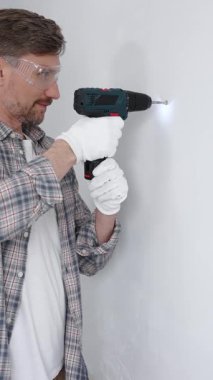 Gri gömlekli ve beyaz eldivenli orta yaşlı işçi, ev yenileme çalışmaları sırasında kablosuz matkapla duvarda delik açıyor. Dikey görünüm.