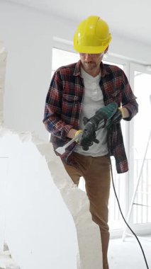 Bej kareli gömlek, sarı başlık ve koruyucu eldiven giyen inşaat işçisi, dönen çekiç matkabıyla beyaz duvarı yıkıyor ve orta uzunlukta toz üretiyor. Yenileme konsepti