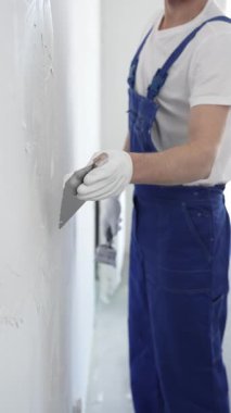 Meçhul inşaat işçisi beyaz eldiven ve mavi tulum giyen kadın, macun bıçağı kullanarak beyaz bir duvara alçı sürüyor. Ev yenileme projesi sırasında pürüzsüz bir yüzey oluşturuyor.