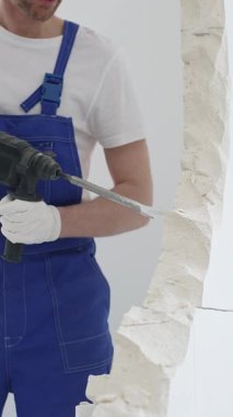 Mavi tulumlu ve koruyucu beyaz eldivenli tanımlanamayan erkek inşaat işçisi, beyaz duvarı döner çekiç matkapıyla yıkıyor, toz üretiyor ve yatay görüşü kapatıyor. Yenileme