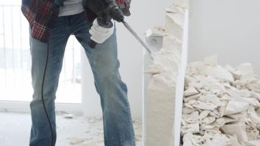 Kırmızı kareli kotlar ve koruyucu eldivenler giyen kimliği belirsiz erkek inşaat işçisi, döner çekiç matkabıyla beyaz duvarı yıkıyor, yerde enkaz oluşturuyor, manzarayı kapatıyor. Yenileme.