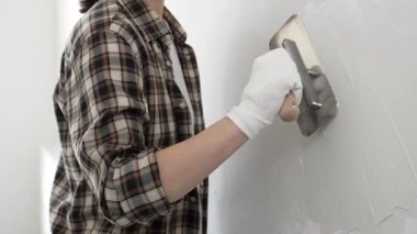 Koruyucu eldiven ve kareli tişört giyen kimliği belirsiz kadın inşaat işçisi, beyaz duvarda ıslak alçıyı düzleştiriyor ve iç dekorasyon sırasında profesyonel malayla malzeme yayıyor.