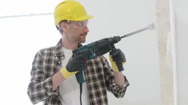 Bej kareli gömlek, sarı başlık ve koruyucu eldiven giyen inşaat işçisi, döner çekiç matkabıyla beyaz duvarı yıkıyor, toz ve portre görüntüsü yaratıyor. Yenileme konsepti.