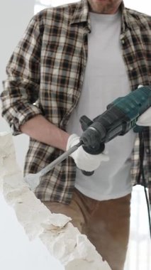 Tanınmayan bej kareli gömlek ve koruyucu eldivenler giyen erkek inşaat işçisi, döner çekiç matkabıyla beyaz duvarı yıkıyor, toz üretiyor, yakın görüş sağlıyor. Yenileme konsepti.