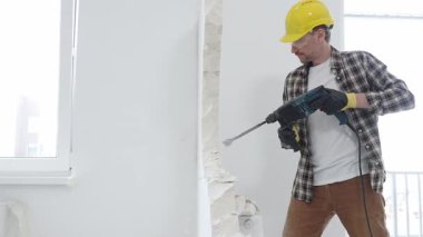Bej kareli gömlek, sarı başlık ve koruyucu eldiven giyen inşaat işçisi, dönen çekiç matkabıyla beyaz duvarı yıkıyor ve orta uzunlukta toz üretiyor. Yenileme konsepti