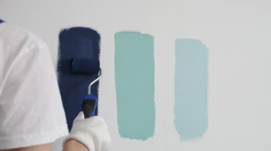 Eldiven giyen ressam beyaz bir duvara boya silindiri uyguluyor, mavi rengin çeşitli tonlarını test ederek iç tasarım için güzel bir eğim etkisi yaratıyor.