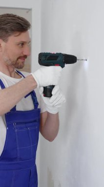 Mavi üniformalı profesyonel işçi, güvenlik eldivenleri, tadilat sırasında beyaz duvarda delik açmak için kablosuz matkap kullanıyor. Dikey portre görünümü.