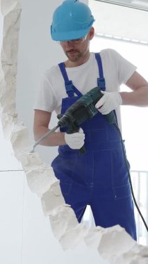 Mavi tulumlu ve koruyucu beyaz eldivenli bir inşaat işçisi, beyaz duvarı döner çekiç matkabıyla yıkıyor, toz üretiyor, yatay görüşü kapatıyor. Yenileme konsepti.