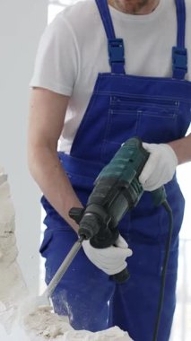 Mavi tulumlu ve koruyucu beyaz eldivenli tanımlanamayan erkek inşaat işçisi, beyaz duvarı döner çekiç matkapıyla yıkıyor, toz üretiyor ve yatay görüşü kapatıyor. Yenileme