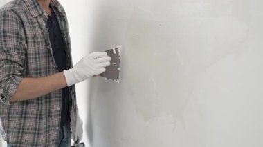 Eldiven ve ekose gömlek giyen tanınmamış inşaat işçisi, bir ev yenileme veya inşaat projesi sırasında beyaz bir duvara sıva uygulamak için macun bıçağı kullanıyor..