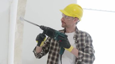 Bej kareli gömlek, sarı başlık ve koruyucu eldiven giyen inşaat işçisi, döner çekiç matkabıyla beyaz duvarı yıkıyor, toz ve portre görüntüsü yaratıyor. Yenileme konsepti.