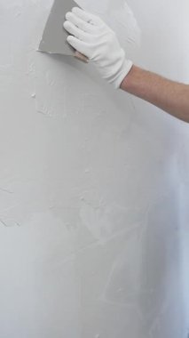 Meçhul inşaat işçisi beyaz eldiven ve mavi tulum giyen kadın, macun bıçağı kullanarak beyaz bir duvara alçı sürüyor. Ev yenileme projesi sırasında pürüzsüz bir yüzey oluşturuyor.