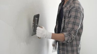Eldiven ve ekose gömlek giyen kimliği belirsiz erkek inşaat işçisi, yenileme projesi sırasında pürüzsüz bir yüzey oluşturmak için beyaz bir duvar alçılamak için son malayı kullanıyor..