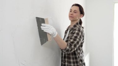 Kareli gömlek giyen kadın inşaat işçisi, beyaz duvar yüzeyini düzleştiriyor, profesyonel spatula tekniği ile macunu uyguluyor, iç dekorasyon konusunda yetenekli..