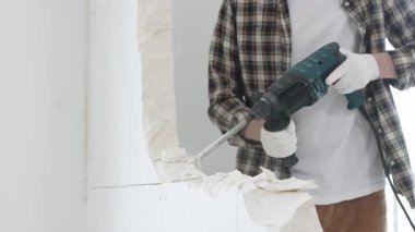 Tanınmayan bej kareli gömlek ve koruyucu eldivenler giyen erkek inşaat işçisi, döner çekiç matkabıyla beyaz duvarı yıkıyor, toz üretiyor, yakın görüş sağlıyor. Yenileme konsepti.
