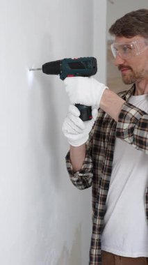 İşaretli gömlek ve beyaz eldiven giyen orta yaşlı bir adam, ev yenileme çalışmaları sırasında kablosuz matkapla duvarda delik açıyor. Dikey görünüm.