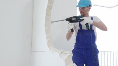 Mavi tulumlu ve koruyucu beyaz eldivenli bir inşaat işçisi, beyaz duvarı döner çekiç matkabıyla yıkıyor, toz üretiyor, yatay görüşü kapatıyor. Yenileme konsepti.