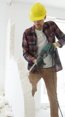 Bej kareli gömlek, sarı başlık ve koruyucu eldiven giyen inşaat işçisi, dönen çekiç matkabıyla beyaz duvarı yıkıyor ve orta uzunlukta toz üretiyor. Yenileme konsepti