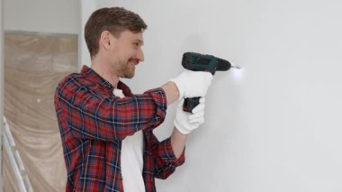 Kırmızı tişört ve beyaz eldiven giyen orta yaşlı erkek işçi, ev yenileme çalışmaları sırasında kablosuz matkapla duvarda delik açıyor. Dikey görünüm.