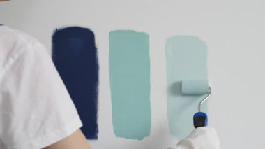 Eldiven giyen ressam beyaz bir duvara boya silindiri uyguluyor, mavi rengin çeşitli tonlarını test ederek iç tasarım için güzel bir eğim etkisi yaratıyor.