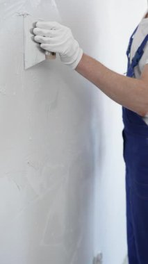 Meçhul inşaat işçisi beyaz eldiven ve mavi tulum giyen kadın, macun bıçağı kullanarak beyaz bir duvara alçı sürüyor. Ev yenileme projesi sırasında pürüzsüz bir yüzey oluşturuyor.