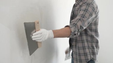 Bilinmeyen erkek inşaat işçisi koruyucu eldivenler ve ekose gömlek giyiyor. Tadilat projesi sırasında profesyonel alçıpan bantlama bıçağını kullanarak duvarın yüzeyine sorunsuzca alçı yayıyor..