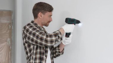 İşaretli gömlek ve beyaz eldiven giyen orta yaşlı bir adam, ev yenileme çalışmaları sırasında kablosuz matkapla duvarda delik açıyor. Dikey görünüm.