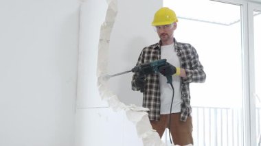 Bej kareli gömlek, sarı başlık ve koruyucu eldiven giyen inşaat işçisi, dönen çekiç matkabıyla beyaz duvarı yıkıyor ve orta uzunlukta toz üretiyor. Yenileme konsepti