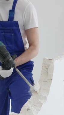 Mavi tulumlu ve koruyucu beyaz eldivenli tanımlanamayan erkek inşaat işçisi, beyaz duvarı döner çekiç matkapıyla yıkıyor, toz üretiyor ve yatay görüşü kapatıyor. Yenileme