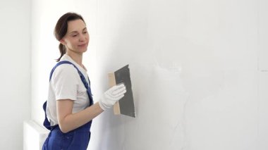 Koruyucu eldivenler ve mavi inşaat tulumları giyen kadın inşaat işçisi, tadilat projesi sırasında profesyonel alçı levha bantlı bıçak kullanarak duvarlara sorunsuzca alçı yayıyor. Portre