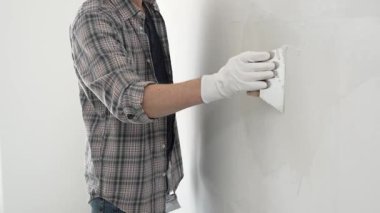 Eldiven ve ekose gömlek giyen tanınmamış inşaat işçisi, bir ev yenileme veya inşaat projesi sırasında beyaz bir duvara sıva uygulamak için macun bıçağı kullanıyor..